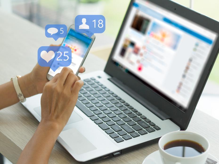 A laptop and a hand scrolling social media on a phone, surrounded by visualisations symbolising likes, comments and followers