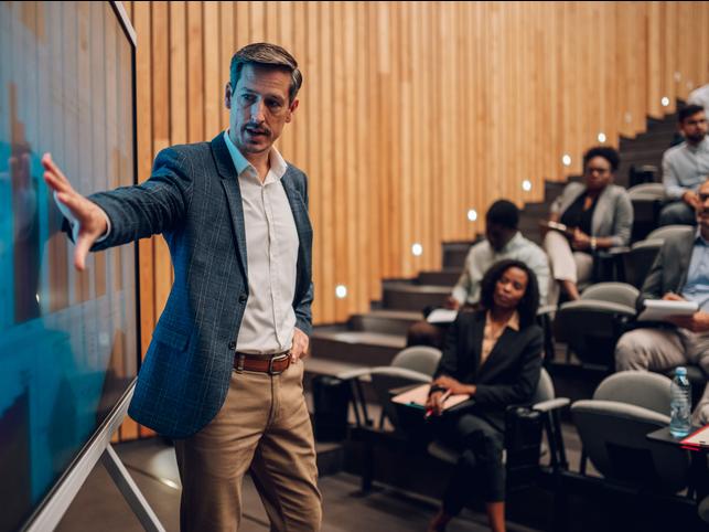 A teacher gestures to a screen in a lecture hall