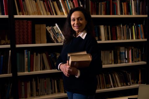 An academic holding a pile of books in front of a library bookshelf