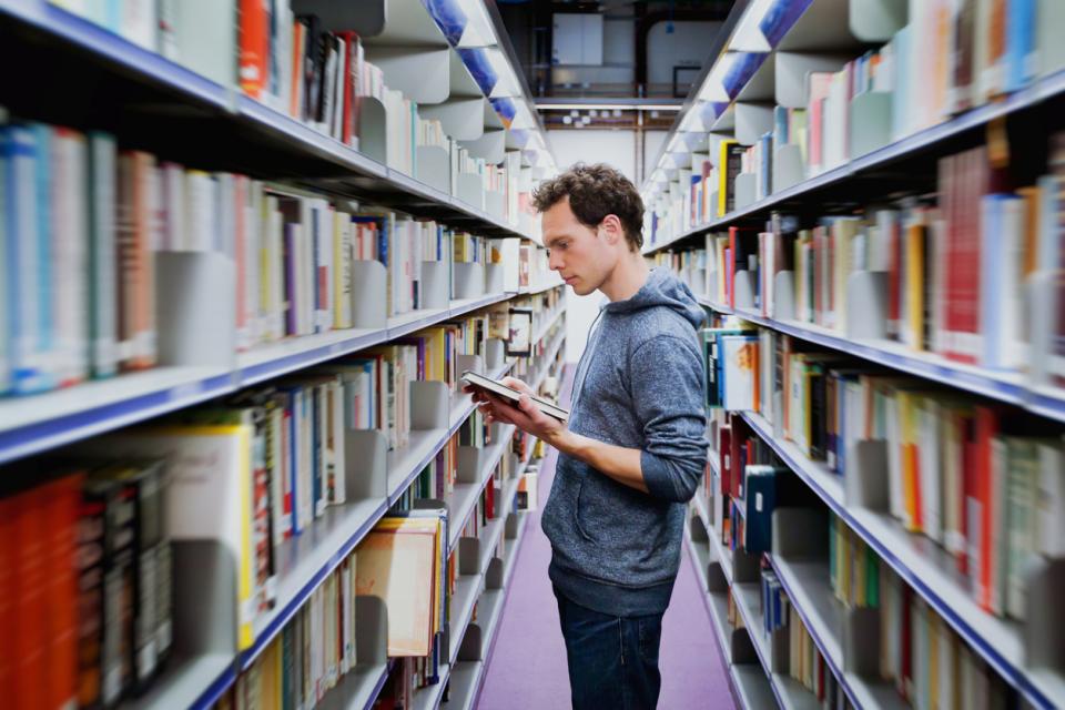 A man reads a book between the aisles of a university library