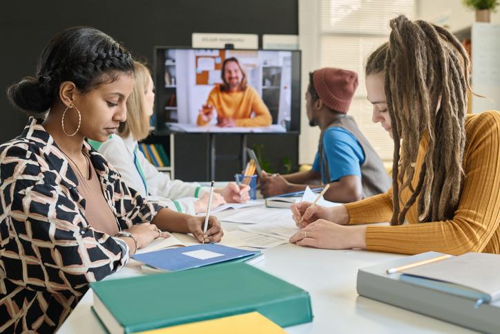 Students in a classroom with an online student appearing on a screen at the end of the table