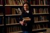 An academic holding a pile of books in front of a library bookshelf